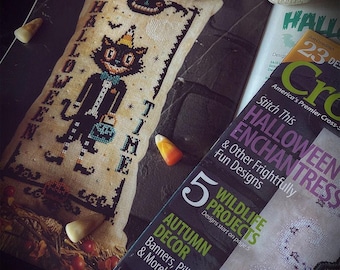 May include: A rectangular cross-stitch sampler featuring a black cat wearing a party hat and holding a basket, with the text HALLOWEEN TIME. The sampler is displayed next to a magazine titled Cross-Stitch with various Halloween-themed text.