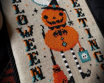 May include: A cross-stitch embroidery on beige fabric featuring a pumpkin character wearing a black hat, holding a broom and a teal bag with a skull. The text HALLOWEEN GREETINGS is stitched in black and teal. The Witchy Stitcher is printed below.