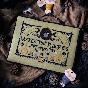 May include: A green cross-stitch sampler featuring the text "Doc Witchcrafts" and various Halloween-themed imagery, including a cauldron, bats, and a crescent moon. The sampler is framed with a black border and has a fringe around the edge.
