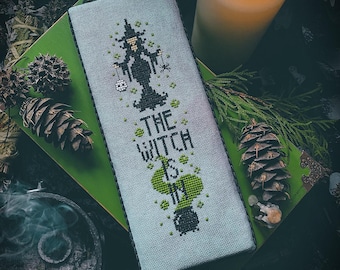 May include: A rectangular cross-stitch sampler on light gray fabric featuring a black silhouette of a witch, a bubbling green cauldron, and the text "THE WITCH IS IN" stitched in black, accented with small green dots and a spider.