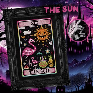 May include: Framed cross-stitch art featuring a flamingo, a jack-o'-lantern sun, and the text "THE SUN". The artwork is set against a black background with a pink border and is displayed in a black ornate frame. The image has a spooky, Halloween theme.