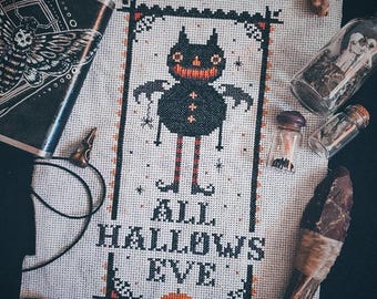 May include: A cross-stitch embroidery on white Aida cloth featuring a black creature with orange accents, surrounded by a decorative border and the text ALL HALLOWS EVE, with a small orange pumpkin at the bottom, displayed among various small trinkets.