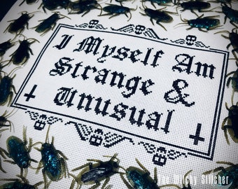 Könnte beinhalten: Eine weiße Kreuzstich-Stickerei mit schwarzem Garn, die den Text "I Myself Am Strange & Unusual" zeigt, umgeben von einem Rand aus schwarzen Schädeln und Schnörkeln. Die Stickerei ist von zahlreichen Plastik-Kakerlaken umgeben.