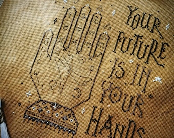 May include: A mustard yellow fabric featuring black cross-stitch embroidery of a palmistry hand chart with zodiac symbols and the text "YOUR FUTURE IS IN YOUR HANDS" and "THE WITCHY STITCHER" stitched in a decorative black font.