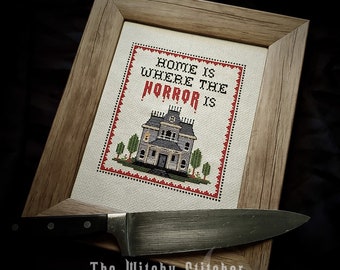 Op de afbeelding: Een ingelijst kruissteekontwerp met een spookhuis en de tekst "Home is where the horror is". De kruissteek is op een crèmekleurige stof met een rode rand. De lijst is van hout en heeft een natuurlijke afwerking. De afbeelding is gesigneerd met "The Witchy Stitcher".