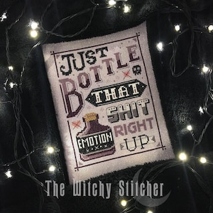 Könnte beinhalten: Eine Kreuzstichstickerei auf hellem Stoff mit einer lila Flaschengrafik und dem Text: JUST BOTTLE THAT SHIT RIGHT UP, wobei ein kleineres Etikett auf der Flasche EMOTION zeigt, umrahmt von einem dunklen Rand und einem kleinen Totenkopfsymbol.
