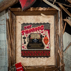 May include: A framed cross-stitch embroidery featuring a black typewriter, a skeleton key, and a red hotel room tag labeled 237. The text reads THE SHINING and all work and no play makes Jack a dull boy. The design uses red, black, and orange thread.