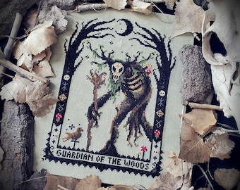 Può includere: Un ricamo a punto croce su tessuto grigio chiaro raffigurante una figura centrale con testa di teschio e corna, che tiene un bastone. Il disegno include alberi, una luna crescente, un piccolo uccello e funghi, con il testo "GUARDIAN OF THE WOODS" in basso.
