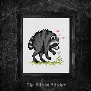 May include: A cross-stitch pattern featuring a pixel art raccoon in shades of gray and black, standing on a patch of green grass. The raccoon is looking at a small blue flower, surrounded by several small pink hearts in a vertical arrangement.