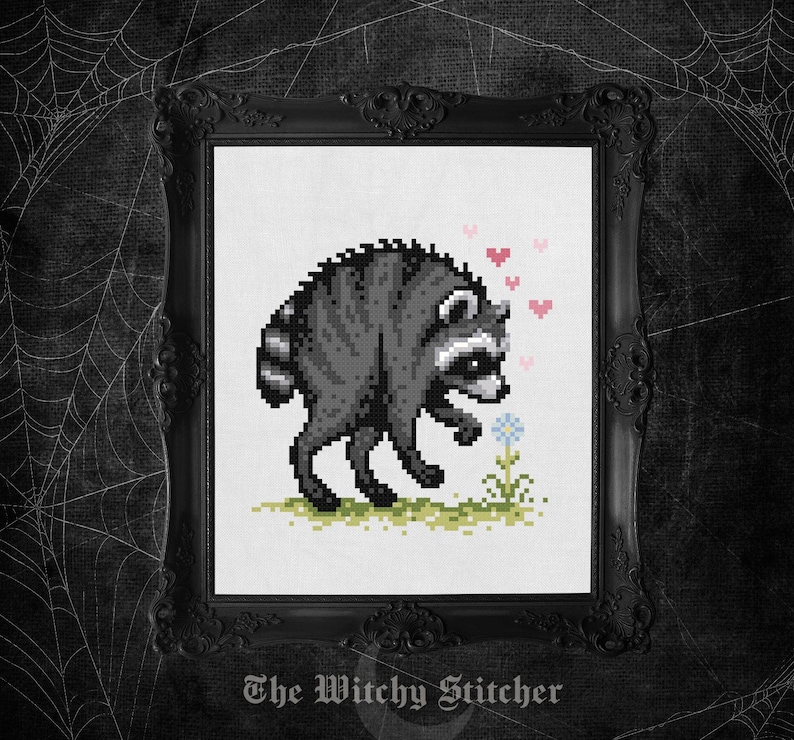 May include: A cross-stitch pattern featuring a pixel art raccoon in shades of gray and black, standing on a patch of green grass. The raccoon is looking at a small blue flower, surrounded by several small pink hearts in a vertical arrangement.