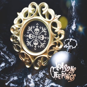 May include: A black and white cross-stitch design of a skull and crossbones in a gold ornate frame. A black Christmas bauble with the text "Seasons Creepings" is next to the frame.