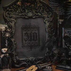 May include: A black ornate frame with a black and white cross-stitch design inside. The cross-stitch design features the word "311" in a gothic font.