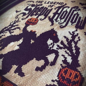 May include: Cross-stitch embroidery of the legend of Sleepy Hollow, featuring a black and white silhouette of the Headless Horseman riding a horse, holding a jack-o-lantern. The background is beige with the text "The Legend of Sleepy Hollow" in black.