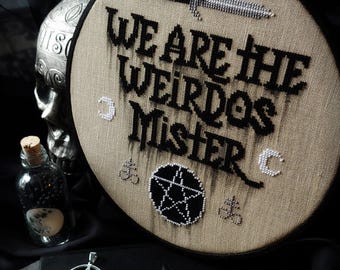 May include: A circular embroidery hoop featuring tan fabric with black cross-stitched text that reads WE ARE THE WEIRDOS MISTER. The design includes a black dagger, a central pentagram, two small crescent moons, and two small alchemical symbols.