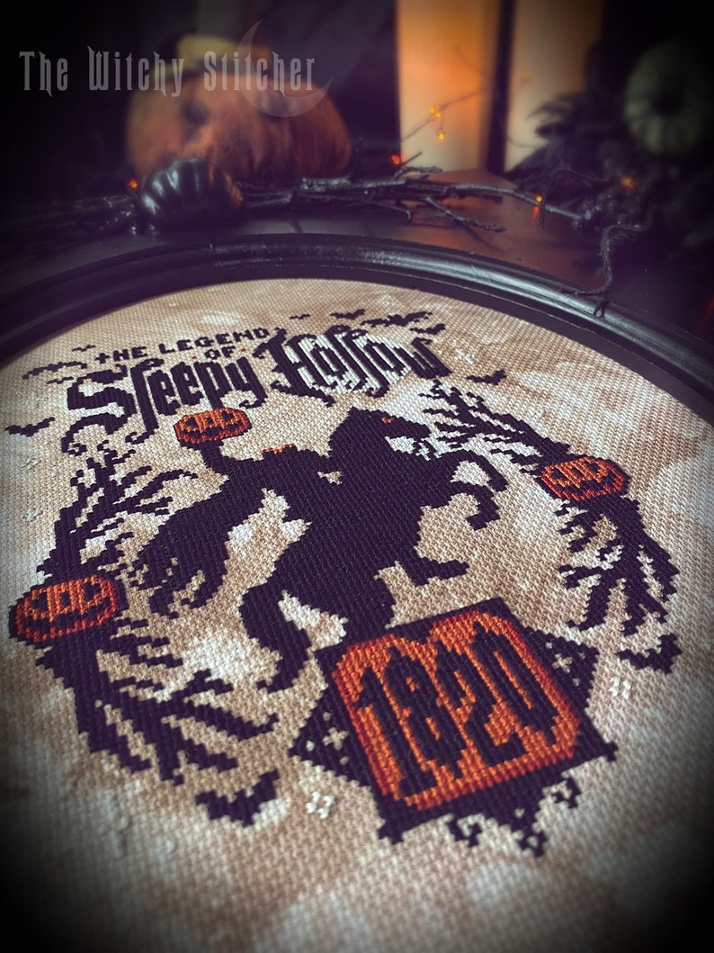 May include: Cross-stitch pattern of a witch riding a broomstick with pumpkins and bats. The text "The Legend of Sleepy Hollow" is above the witch. The year "1920" is in the bottom right corner.