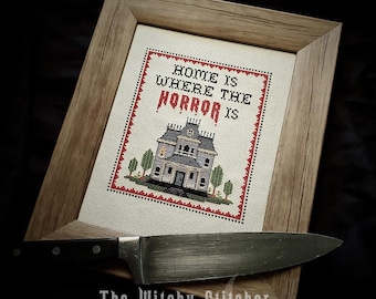 May include: A framed cross-stitch embroidery featuring a haunted house illustration and the text "HOME IS WHERE THE HORROR IS" in black and red thread, accented by a red and black border, with a large kitchen knife resting across the frame.