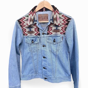 Könnte beinhalten: Hellblaue Jeansjacke mit gemustertem Kragen und Schulterpasse. Die Passe hat ein geometrisches Design in Rot-, Braun- und Blautönen. Die Jacke hat eine Knopfleiste und ein kleines Etikett mit der Aufschrift "EDWIN".