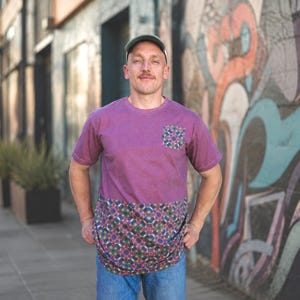 Könnte beinhalten: Ein lila T-Shirt mit einer gemusterten Tasche und einem unteren Panel. Die Tasche und das untere Panel weisen ein sich wiederholendes geometrisches Design in Blau-, Grün- und Rosatönen auf. Das Shirt wird mit einer blauen Jeans kombiniert.