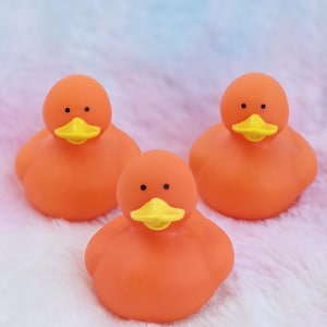 Peut inclure: Trois canards en caoutchouc orange avec des becs jaunes et des yeux noirs sont disposés sur une surface douce et multicolore. Les canards sont un jouet de bain classique, parfait pour les enfants.