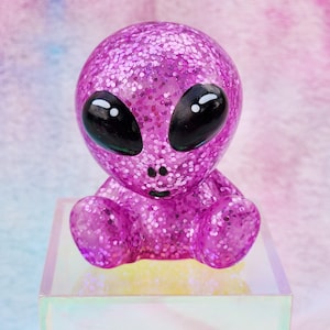 May include: A small, pink, glitter-covered alien figurine with black eyes and a simple smile. The figurine is sitting on an iridescent, square base.