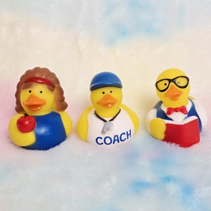 May include: Three yellow rubber duckies, each with unique accessories. One duck wears a headband and holds a red apple. Another is dressed as a coach with a whistle. The third duck wears glasses and a bow tie, holding a red book.