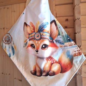 Peut inclure: Une couverture pour bébé avec un renard de dessin animé portant une coiffe amérindienne. Le renard est orange et blanc avec de grands yeux. La couverture a un fond crème avec des plumes colorées et des motifs géométriques.