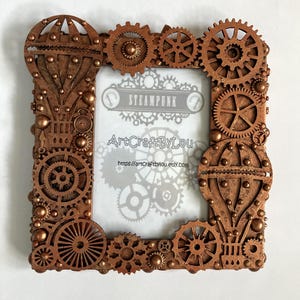 May include: A copper-coloured picture frame decorated with gears, hot air balloons, and rivets. The frame has a white background with the text "STEAMPUNK" and "ArtCraftByLou" in black.