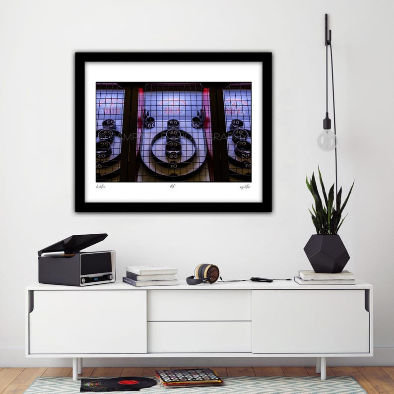 May include: A framed photograph of a skee-ball machine with a black frame. The machine has a series of circular targets with point values. The background is a grid of lights. The photograph is displayed on a white cabinet with a record player and a plant.