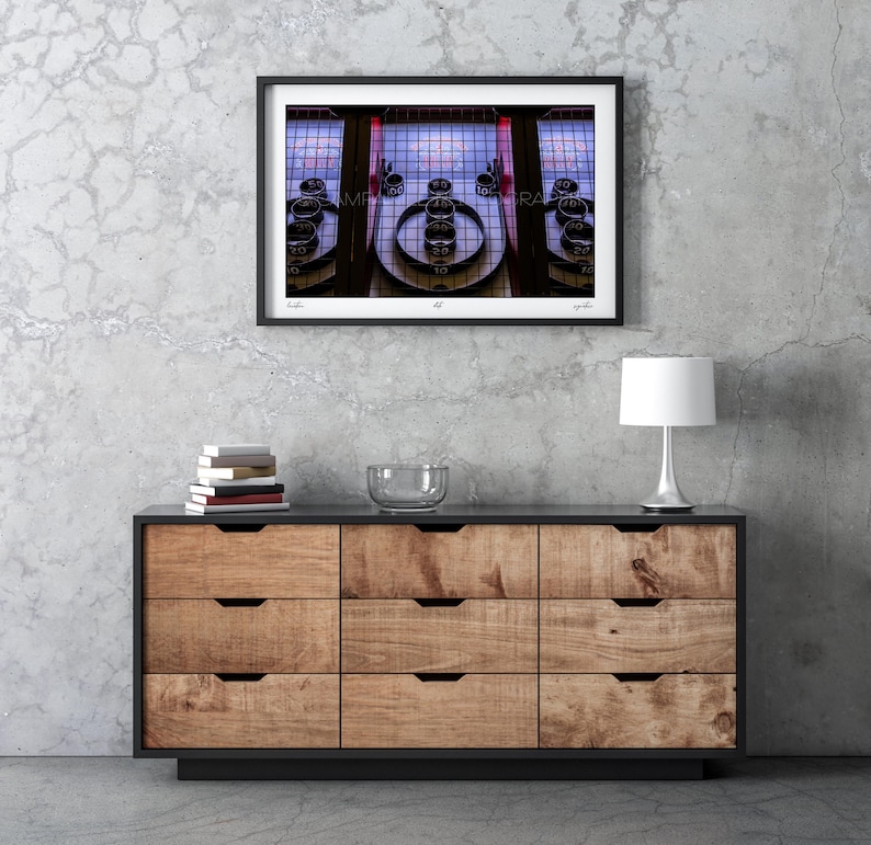 May include: A framed photograph of a vintage Skee-Ball machine. The image is in a black frame and hangs above a wooden dresser. The Skee-Ball machine has a series of numbered targets and a red and blue color scheme.
