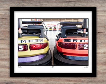May include: A framed print featuring two bumper cars, one yellow with the number 5 and the other red with the number 2. The bumper cars are positioned close together, with a blurred background of other bumper cars.