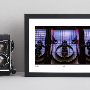 May include: Framed photograph of a Skee-Ball machine, a classic arcade game. The image captures the game's layout, with ramps, scoring rings, and numbered targets. The photograph is in a black frame, with a vintage camera on the left.