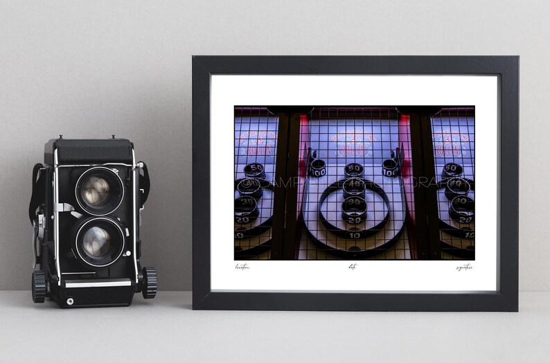 May include: Framed photograph of a Skee-Ball machine, a classic arcade game. The image captures the game's layout, with ramps, scoring rings, and numbered targets. The photograph is in a black frame, with a vintage camera on the left.