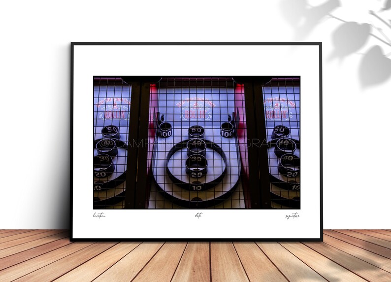 May include: A framed print of a vintage Skee-Ball arcade game. The game features numbered targets and ball return lanes. The print is displayed on a wooden surface.
