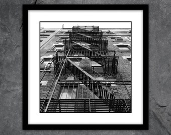 May include: Black and white photograph of a fire escape on the side of a brick building. The fire escape is made of metal and has several levels. The photo is taken from a low angle, looking up at the fire escape.