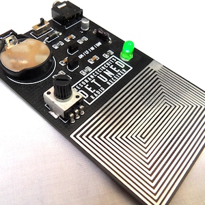 Puede incluir: Una placa de circuito impreso negra con una luz LED verde, una perilla plateada y una batería. La placa tiene un patrón de circuito impreso y el texto "RESONANCE CIRCUITS DETUNED RADIO RECEIVER".