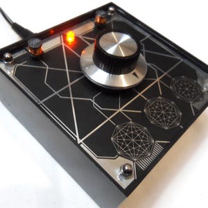 Può includere: Una scatola nera con una manopola argentata e una luce LED rossa. La scatola ha un motivo geometrico sulla parte superiore.