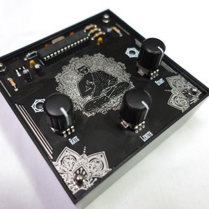 Puede incluir: Un pedal de efectos de música electrónica negro con tres botones etiquetados "Rate", "Length" y "Shift". El pedal presenta un diseño de mandala plateado y un gráfico de una figura meditando.