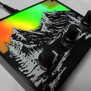 Puede incluir: Dispositivo electrónico negro con un diseño de cordillera. El dispositivo tiene tres perillas negras con indicadores blancos y un fondo degradado colorido de naranja, verde y amarillo. Un cable negro está enchufado.