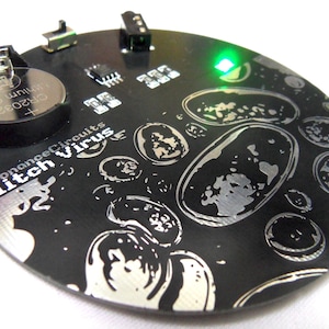Puede incluir: Una placa de circuito circular negra con un diseño plateado de burbujas y el texto "Resonance Circuits Glitch Virus". Una luz LED verde está encendida en la esquina superior derecha de la placa.