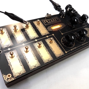 May include: A black electronic sound synthesizer labeled Resonance Circuits Entropy Generator with gold-colored rectangular touch plates, black rotary knobs, a glowing orange light display, and a black alligator clip lead attached to one plate.