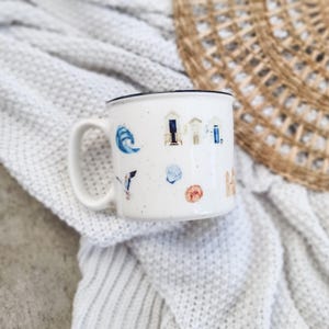 May include: White ceramic mug with a blue rim and a speckled design. The mug features illustrations of beach houses, seashells, a wave, and a seagull.