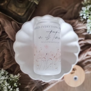 May include: A frosted glass can-shaped tumbler with the text "Makes Everything Beautiful in its Time" and "Ecclesiastes 3:11". The tumbler features a delicate floral design in shades of pink and brown. It sits on a white scalloped plate.