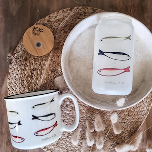 May include: A white ceramic mug and a glass tumbler with fish illustrations in green, black, and red. The mug has a black rim and a handle. The tumbler has a wooden lid. The text "Matthew 4:19" is on both items.