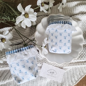 May include: Two mason jars wrapped in white fabric with blue floral patterns and trimmed with blue gingham and white lace. The jars sit on a white scalloped plate and a white knitted blanket. White flowers are arranged around the jars.