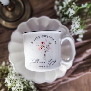 May include: White ceramic mug with black rim, floral design, and the text "IN YOUR PRESENCE IS fullness of joy" with "PSALM 16:11" below. The mug sits on a white scalloped plate, with a candle holder and flowers in the background.