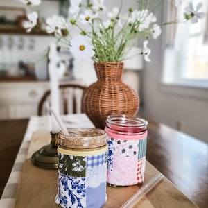 May include: Two glass jars with fabric sleeves, one with a straw. The sleeves are patchwork with floral and patterned fabrics. A clear straw rests on the wooden surface. A vase with white flowers sits in the background.