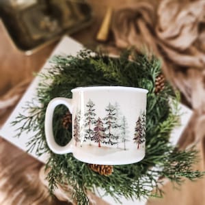 May include: A white ceramic mug with a forest scene design featuring green, red, and gray trees. The mug has a large handle and is surrounded by a green wreath with pinecones. The mug is on a white square plate.