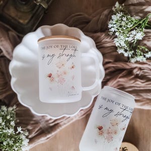 May include: Frosted glass mug and can-shaped glass with floral design and the text "THE JOY OF THE LORD is my strength." The mug has a handle and a wooden lid. The can-shaped glass has a wooden lid. Both are on a white plate.