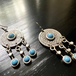 May include: Silver dangle earrings with a round, ornate design and blue stones. The earrings have a floral pattern and multiple dangling stones.