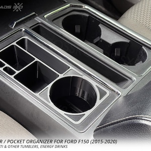 May include: A black cup holder and pocket organizer for a Ford F150 (2015-2020). The organizer has multiple compartments and a cup holder. The text reads: CUP HOLDER / POCKET ORGANIZER FOR FORD F150 (2015-2020).